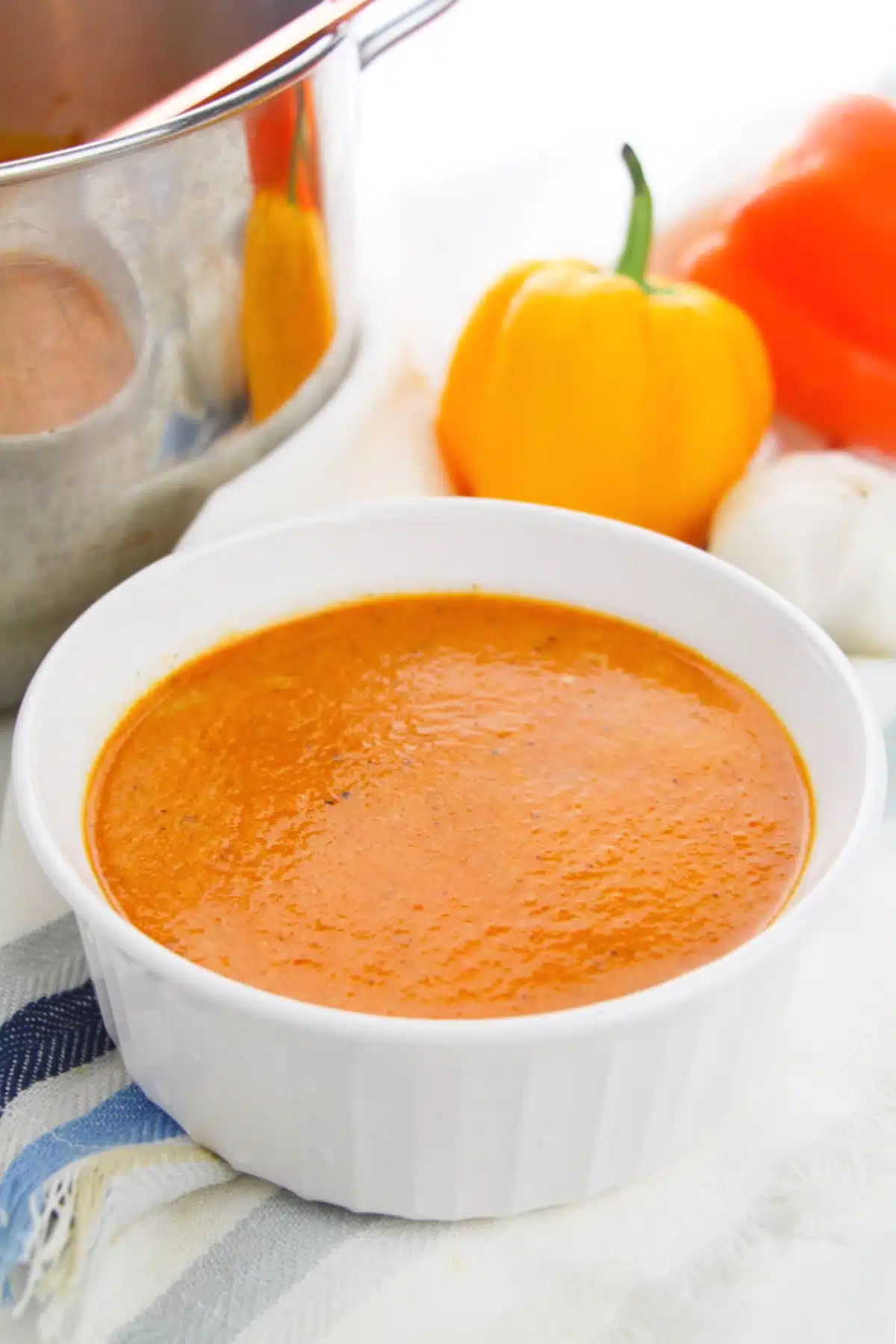 Roasted pepper soup is served in a bright white bowl. Whole bell peppers and a garlic bulb are in the background, and the stainless pot of soup sits to the side.