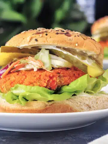 A buffalo cauliflower burger in a bun sits on a white plate. The burger is dressed with butter lettuce, slaw, pickle slices, mayonnaise and a seeded bun.