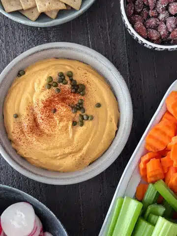 A bowl of roasted pepper dip garnished with capers and paprika spice sits on a wood surface. Served with the dip is a platter of fresh veggies like sliced carrots , celery, and peppers, a bowl of almonds, a bowl of sliced radish, and a bowl of crackers.
